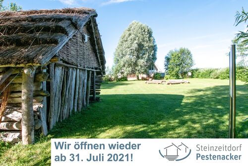 Im Vordergrund das Steinzeithaus und im Hintergrund die Feuerstelle und der Besucherpavillon